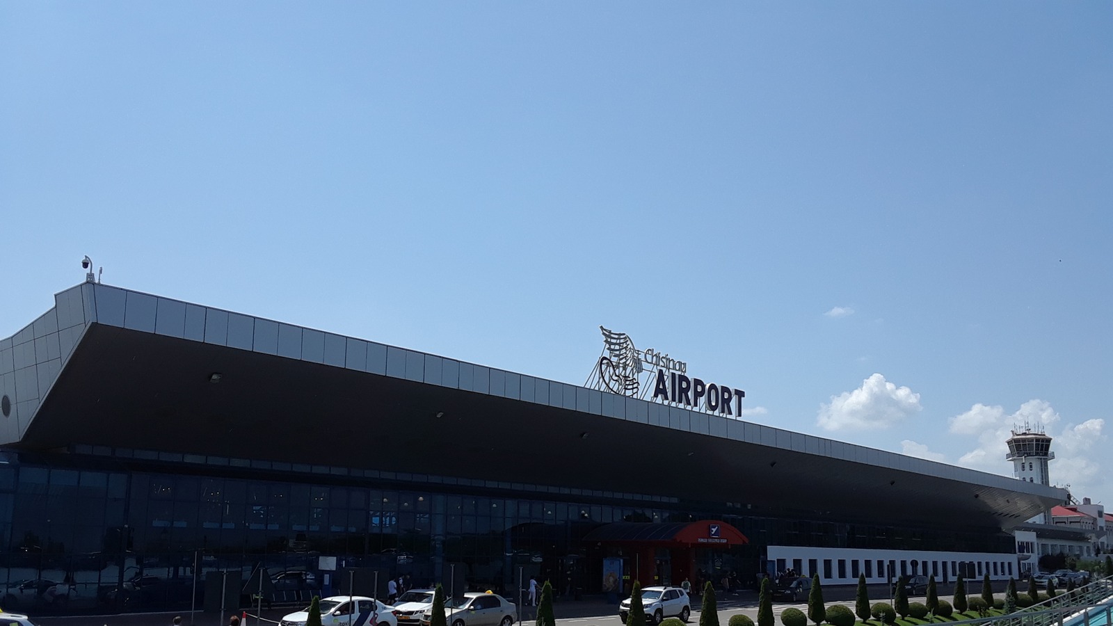 Exterior of Chisinau International Airport terminal with airport signage, glass facade, control tower, and curbside vehicles under a clear sky.