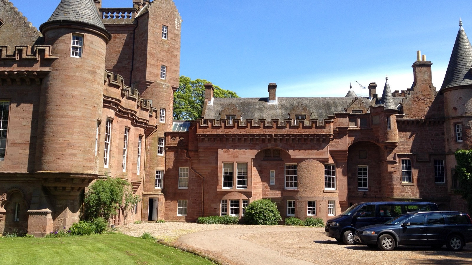 Hospitalfield House in Arbroath with stone towers, a gravel drive, and two parked cars beneath a blue sky.