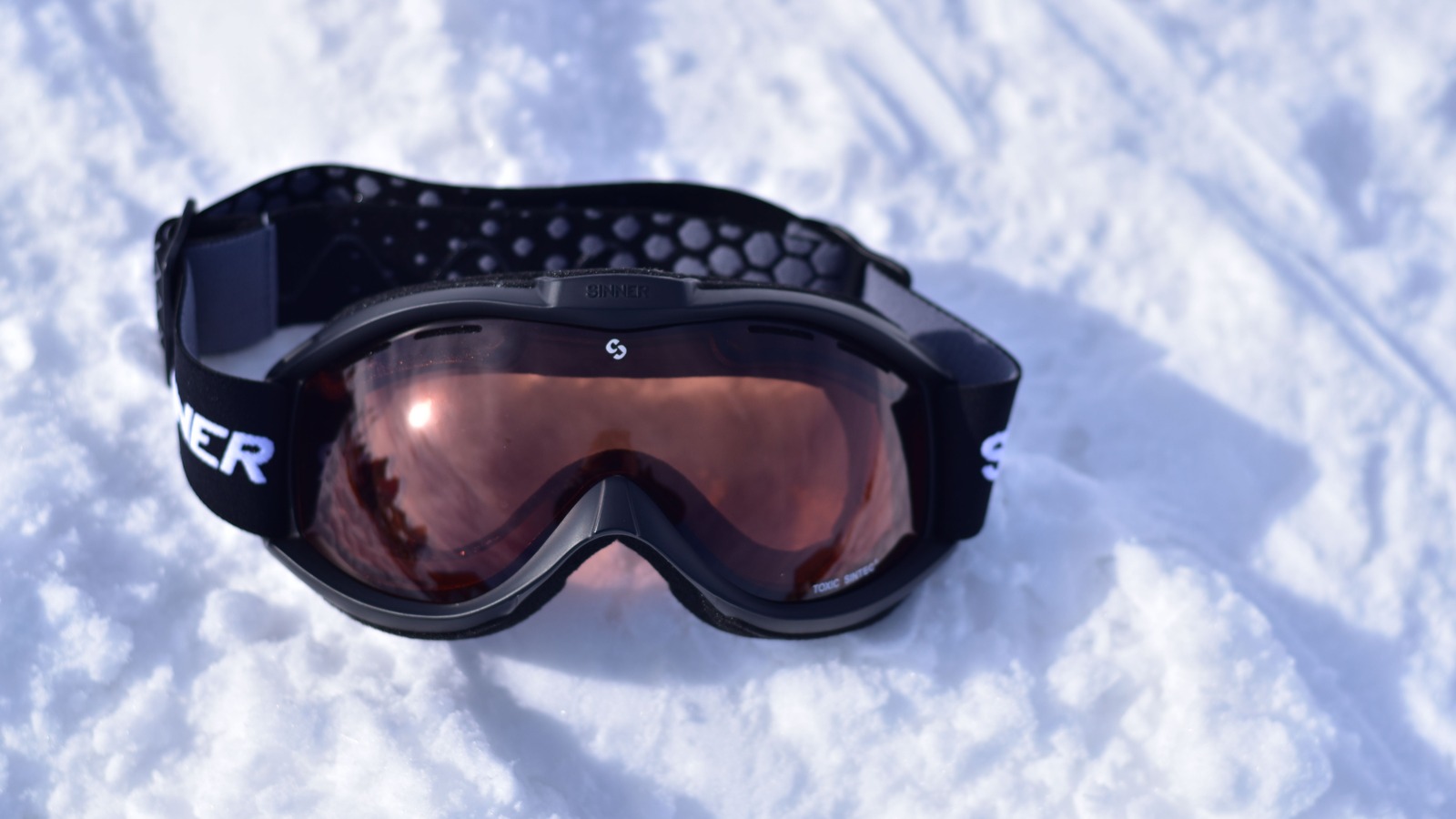 A pair of snowboard goggles resting on snow, with a broad reflective lens.