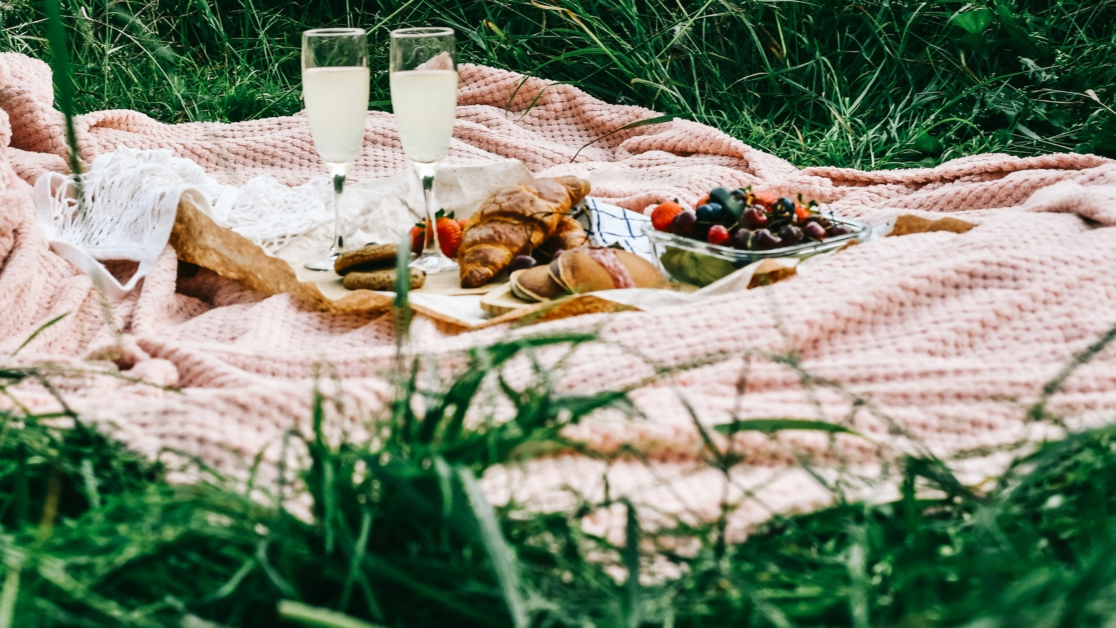 A pale woven picnic blanket with drinks and food spread across grass.