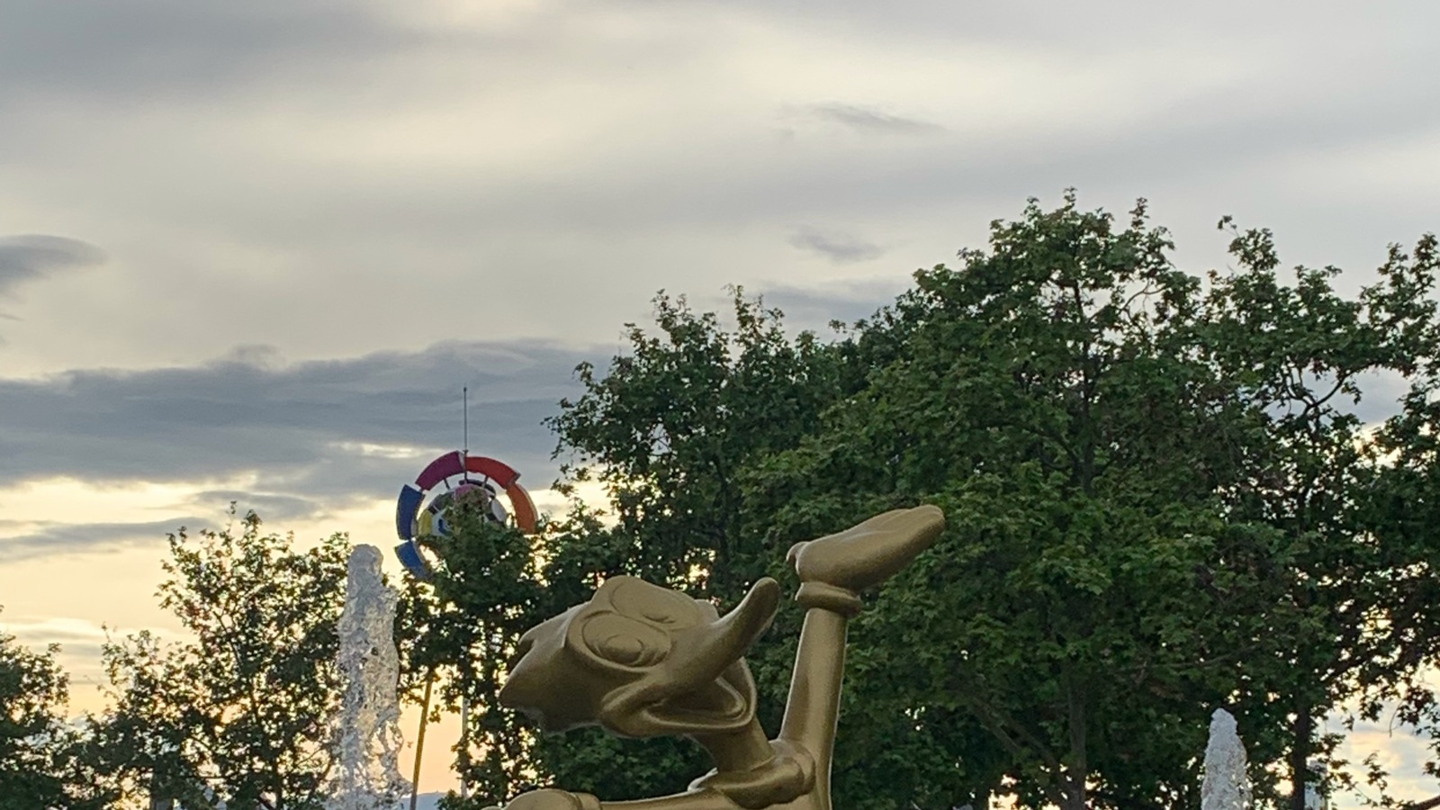 A gold Woody character stands above a fountain, with part of a colorful entrance arch visible behind trees at PortAventura Park.