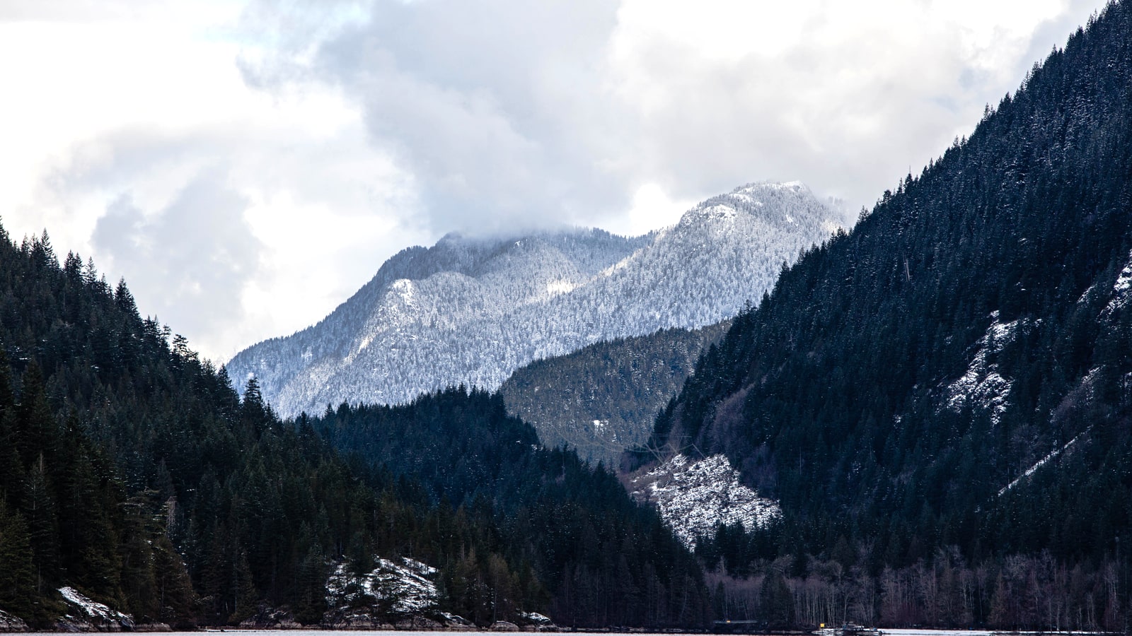 Snow dusted mountains and dark forested ridges rise above the edge of Buntzen Lake.